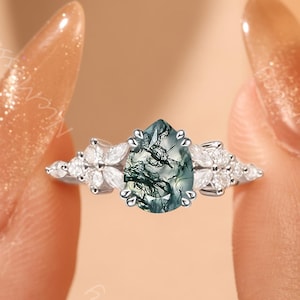 May include: A silver ring featuring a pear-shaped, moss agate gemstone with green and white patterns. The ring is flanked by small, white, marquise-cut stones on each side, creating a floral design. The ring is held between two fingers.