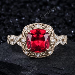 May include: A gold ring featuring a large, cushion-cut red gemstone at the center, surrounded by an intricate filigree frame set with small white stones. The band has a twisted, interwoven design adorned with tiny white stones along the sides.