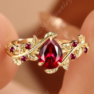 May include: A gold ring with a central, pear-cut, deep red gemstone. The ring is decorated with leaf and berry motifs. The image includes the text "NicMossJewelry".