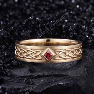 May include: A gold band ring featuring an intricate Celtic knot pattern engraved along the circumference. A single square-cut red gemstone is set in a diamond-shaped frame at the center of the band, creating a focal point on the metal surface.