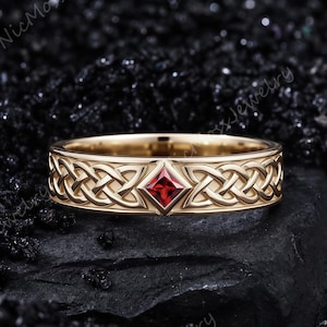 Puede incluir: Un anillo de banda de oro con un intrincado patrón de nudos entrelazados a lo largo de toda la circunferencia. Una piedra preciosa roja de corte cuadrado está engastada en un marco en forma de diamante en el centro. Sobre una superficie oscura.