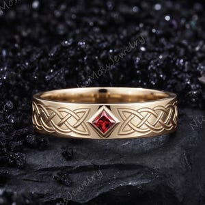 Puede incluir: Un anillo de banda de oro con un intrincado patrón de nudos celtas grabado alrededor del exterior. Una piedra preciosa roja de corte cuadrado está engastada en un marco en forma de diamante en el centro, contrastando con el metal dorado.