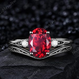 May include: A silver ring set with a large, oval-cut, deep red gemstone. The ring is accented with two smaller, clear gemstones. The band features an ornate filigree pattern. The ring is displayed on a dark, textured surface.