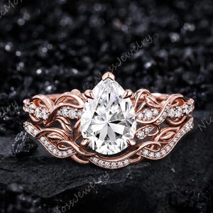 May include: A rose gold ring featuring a large pear-shaped center stone surrounded by a band with intricate vine-like metalwork and small pavé-set stones. The design includes two bands that appear to be joined, creating a layered, botanical look.