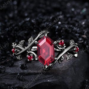 May include: A black metal ring featuring a large, faceted red gemstone at the center. The ring is adorned with a floral design, with smaller red gemstones and leaf accents. The ring is set against a dark, textured background.