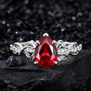 May include: A silver ring featuring a large pear-cut red gemstone at the center, held by prongs. The band is designed with intricate vine-like metalwork and small, clear round stones embedded along the sides, creating a detailed, nature-inspired look.