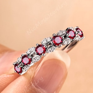 May include: A silver ring featuring a row of vibrant red gemstones, interspersed with smaller, clear stones. The ring is held against a neutral background, showcasing its delicate design and sparkling details. The ring is a piece of jewelry.