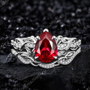May include: A silver-toned ring featuring a large, pear-shaped, deep red gemstone at the center. The band is crafted with intricate, vine-like metalwork and is accented with numerous small, clear, sparkling stones along the detailed branches.