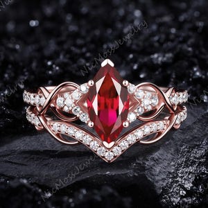 May include: A rose gold ring set featuring a marquise-cut deep red center stone surrounded by small clear accent stones. The band has an intricate, intertwined design with additional clear stones embedded along the metal surface.