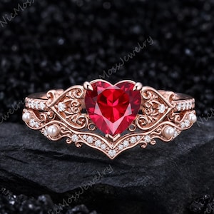 Puede incluir: Un anillo de oro rosa con una piedra preciosa roja central en forma de corazón, rodeada de intrincados filigranas de metal, pequeñas perlas blancas y diminutas piedras claras a lo largo de la banda, con el texto NicMossJewelry en el fondo.