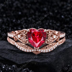 Puede incluir: Un anillo de oro rosa con una piedra preciosa roja central en forma de corazón. La banda y el engaste están decorados con intrincados filigranas de metal y pequeñas piedras blancas. El diseño incluye una banda curva y ornamentada.