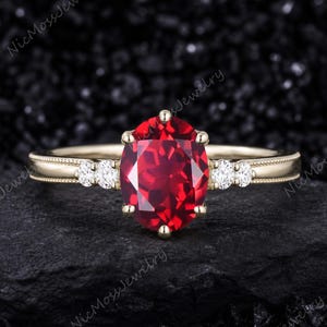 May include: A gold ring featuring a large oval-cut red gemstone at the center, held by six prongs. The band is accented with two small round white stones on each side and features a delicate beaded milgrain edge along the metal shank.