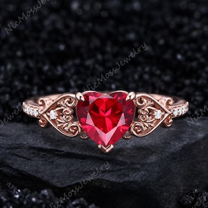 May include: A rose gold ring featuring a central heart-shaped red gemstone. The band displays intricate scrollwork patterns on both sides of the center stone, accented with small clear round stones embedded along the shank of the metal band.
