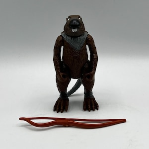 May include: A brown action figure of a beaver-like character with a gray scarf and wristbands. The figure has a black nose and a long gray tail. A red bow is in front of the figure.