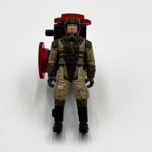 May include: A vintage action figure of a military-style character in a tan uniform with black boots and gloves. The figure has a helmet and a red and black accessory attached to its back. The figure is standing on a white surface.
