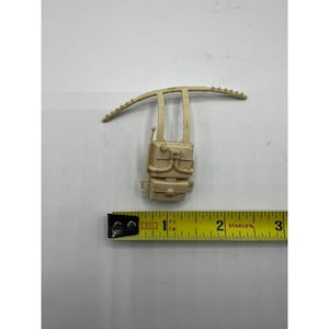 May include: A tan-colored, plastic toy backpack with a curved top and a rectangular body. The backpack has several compartments and details, including a small antenna. A yellow measuring tape is placed next to the backpack.