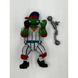Puede incluir: Figura de acción de las Tortugas Ninja en uniforme de béisbol. La figura es verde con una máscara roja y una gorra de béisbol azul. El uniforme es blanco con rayas azules y detalles rojos. Un arma plateada está al lado de la figura.