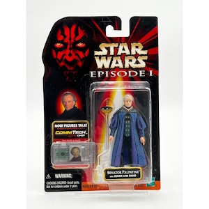 Könnte beinhalten: Star Wars Episode I Actionfigur von Senator Palpatine mit Senate Cam Droid. Die Figur befindet sich in einer transparenten Kunststoffhülle, die an einer schwarz-roten Karte mit dem Text "NOW FIGURES TALK!" und "COMMTECH CHIP" befestigt ist.