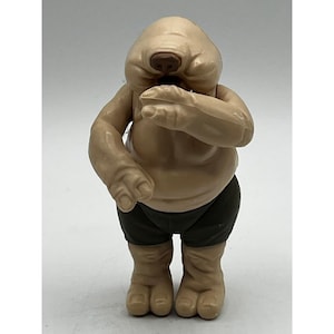 Può includere: Una statuetta colorata color camoscio di una creatura umanoide con una testa grande, piccoli occhi e una bocca larga. La statuetta indossa pantaloncini verde scuro. La statuetta è in piedi con le braccia piegate e le mani vicino al viso.