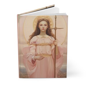 May include: A pink and gold illustrated journal cover featuring a woman with long brown hair holding a spear with a cross on top. The woman is wearing a pink dress with gold accents and a halo.