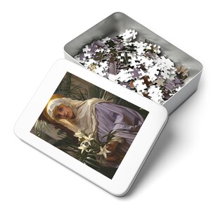 May include: A white metal tin with a white jigsaw puzzle inside. The puzzle depicts a woman in a white and purple robe holding a bouquet of white lilies.