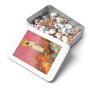 May include: A white tin box containing a colourful jigsaw puzzle. The puzzle shows a painting of a man in white robes standing in a field of fire. The man has a halo around his head.