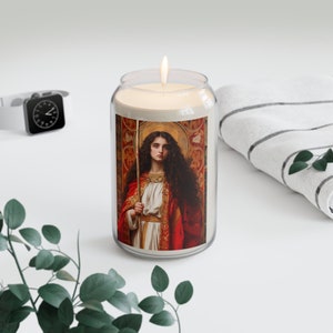 Op de afbeelding: Een glazen kaarsglas met een witte kaars erin. De pot heeft een etiket met een schilderij van een vrouw in een rode en gouden jurk die een staf vasthoudt. De vrouw heeft lang bruin haar en kijkt naar rechts.