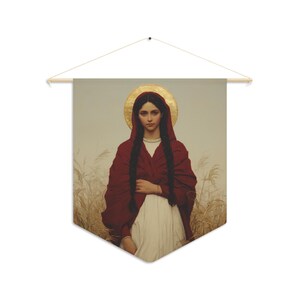 May include: A wall hanging banner featuring a portrait of a figure with long dark braided hair, wearing a deep red hooded cloak over a white garment. A golden circular halo is positioned behind the head, set against a field of tall dry grass.