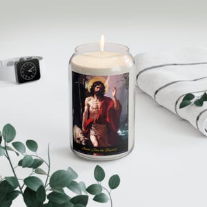 Puede incluir: Una vela encendida en un tarro de cristal transparente con una etiqueta que presenta una pintura de San Juan Bautista. La pintura representa a un hombre con una túnica roja y un halo. El texto "Saint John the Baptist" está impreso en la etiqueta.
