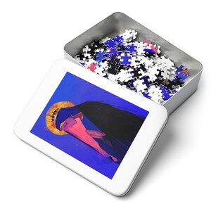 May include: A white metal tin with a blue, pink, and gold illustration of a woman wearing a crown and a blue robe. The tin is open and filled with a jigsaw puzzle.