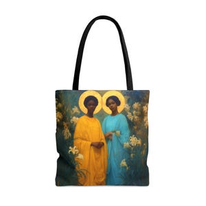 May include: Tote bag featuring a painting of two women with halos, dressed in yellow and blue robes, standing in a garden of white lilies. The bag has black handles and a colorful, detailed print.