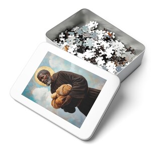 May include: A white metal tin with a lid open, revealing a completed jigsaw puzzle. The puzzle depicts a painting of a man in a brown robe holding loaves of bread. The man has a halo above his head.