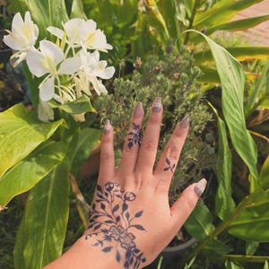 May include: A hand with a dark blue floral tattoo on the wrist and fingers. The nails are painted with a neutral colour and a plaid design. The background features green foliage and white flowers.