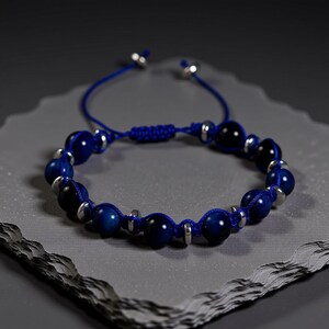 May include: A blue beaded bracelet with silver-colored accents. The bracelet features dark blue, spherical beads strung on a blue cord. The cord is adjustable with two silver-colored beads at the ends. The bracelet is displayed on a gray surface.