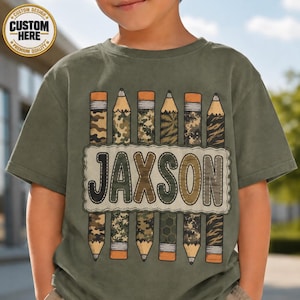 May include: An olive green t-shirt featuring a graphic of six vertical pencils with various camouflage patterns. A central cream-colored rectangular patch with a scalloped border displays the name JAXSON in large, dark brown, stitched-style letters.