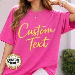May include: A vibrant pink t-shirt with the words "Custom Text" in a glittery gold script. The shirt also features a circular emblem that reads "Custom Design" and "Custom Here". The shirt is a casual style.