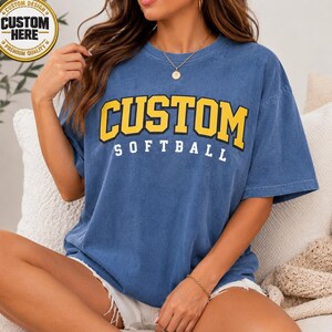 May include: Blue t-shirt with the word "CUSTOM" in yellow block letters with a black outline, above the word "SOFTBALL" in white. The shirt has a vintage, washed look. A gold emblem is in the upper left corner.