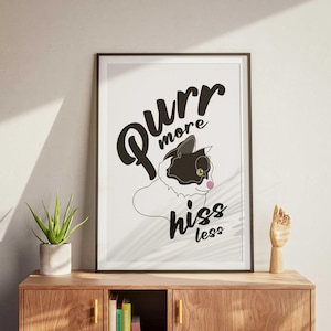 Pode incluir: Impressão artística emoldurada com uma ilustração de gato a preto e branco e o texto "Purr more hiss less". O gato tem um nariz rosa e um olho verde. A impressão está numa moldura preta e é exibida numa cómoda de madeira.