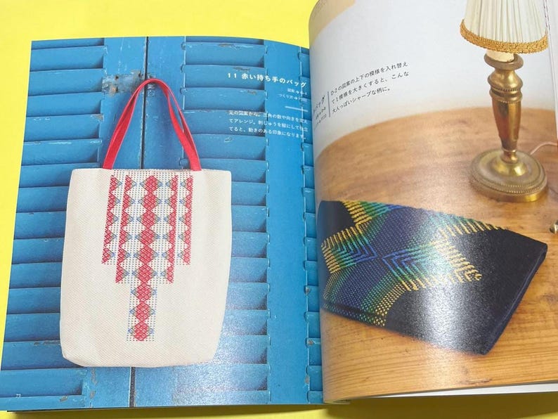 Puede incluir: Un bolso tote blanco con asas rojas y un patr&oacute;n geom&eacute;trico en rojo y azul claro. El bolso se muestra sobre un fondo azul, lo que sugiere un art&iacute;culo artesanal o hecho a mano.
