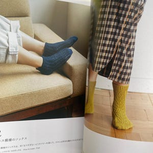 May include: A pair of dark blue ankle socks with a textured knit pattern and a pair of mustard yellow mid-calf socks featuring a vertical cable knit design. The socks are displayed on feet within a printed page showing knitting instructions.