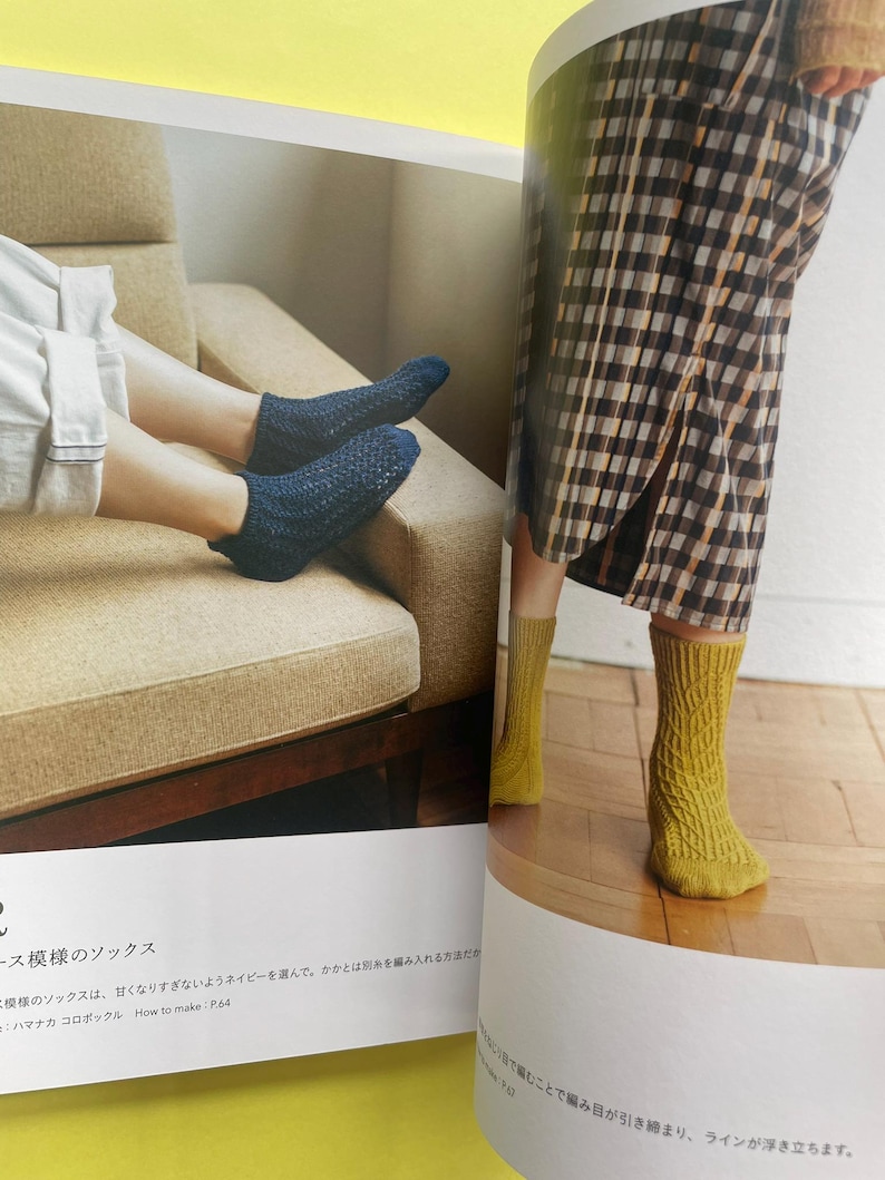 May include: A pair of dark blue ankle socks with a textured knit pattern and a pair of mustard yellow mid-calf socks featuring a vertical cable knit design. The socks are displayed on feet within a printed page showing knitting instructions.