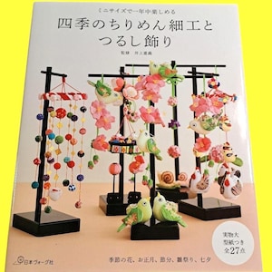 May include: A book cover featuring Japanese craft projects. The title reads "Shiki no Chirimen Saiku to Tsurushi Kazari" with Japanese text and illustrations of hanging ornaments, birds, and flowers. The book is on a yellow background.