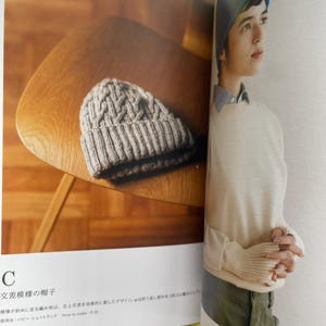 May include: A beige knit beanie with a textured cable pattern and a ribbed cuff. The page includes the text C, 交差模様の帽子, and How to make: P.36. The beanie is displayed on a wooden surface next to a photograph of a person wearing a blue beanie.