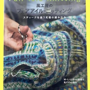 May include: A book cover titled "Fair Isle Knitting" in English and Japanese. The cover features a close-up of a knitted sweater in shades of blue, green, and yellow. Scissors are cutting the knitted fabric. The book's title and author are visible.
