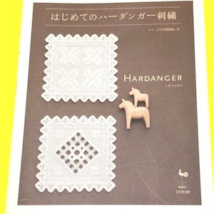 Op de afbeelding: Boekomslag met de titel "HARDANGER" en Japanse tekst. Het toont twee witte, vierkante, geborduurde stukken en twee kleine houten paardenbeeldjes. De achtergrond is bruin.