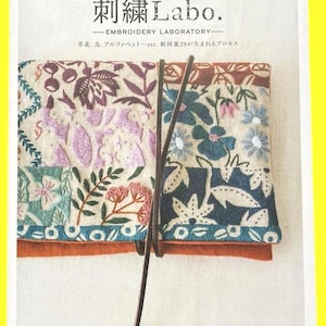 May include: A book cover with the title "Embroidery Labo" in Japanese. The cover features a colorful, embroidered design with floral and leaf patterns in shades of purple, blue, and orange. A leather strap wraps around the book.