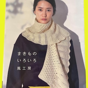 以下が含まれることがあります： クリーム色のオープンニットのスカーフを着用した女性。スカーフはフリルエッジで、濃い色のトップスにドレープされています。画像には、本に日本語のテキストが表示されています。