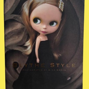 May include: A book cover featuring a doll with large green eyes, wearing a black dress and a jeweled hair clip. The title "BLYTHE STYLE" is displayed in gold lettering, with the author's name below.