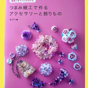 May include: A pink book cover with Japanese text and various handmade fabric flower accessories. The accessories include a butterfly, floral wreaths, and individual flowers in shades of purple, pink, and blue. The book title is "Accessories and Decorations Made with Tsumami Craft."