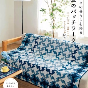 以下が含まれることがあります： ベージュのソファを覆う青と白の模様のキルト。キルトは、さまざまな青の色合いの様式化された花の形の繰り返しデザインが特徴です。表紙のテキストは「日本の青いパッチワーク」と書かれています。サイドテーブルにはお揃いのプレースマットとティーカップがあります。